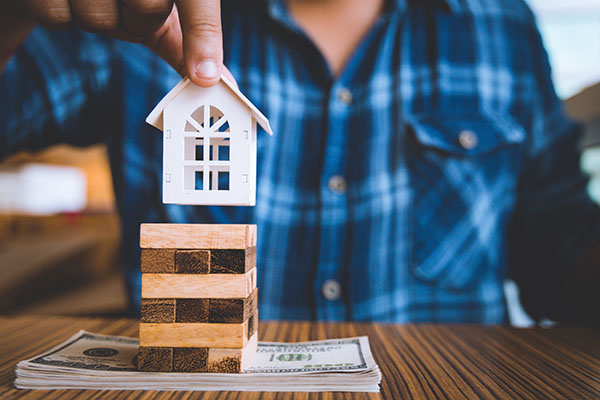 A person placing a model house on top of stack of block which is on top of a stack of paper money