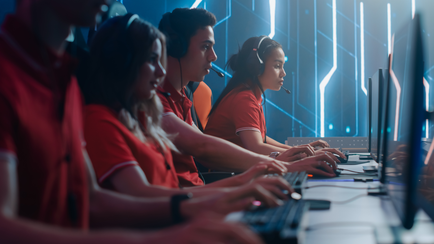 Four people sit in front of four computers in a row in a gaming room and play computer games.